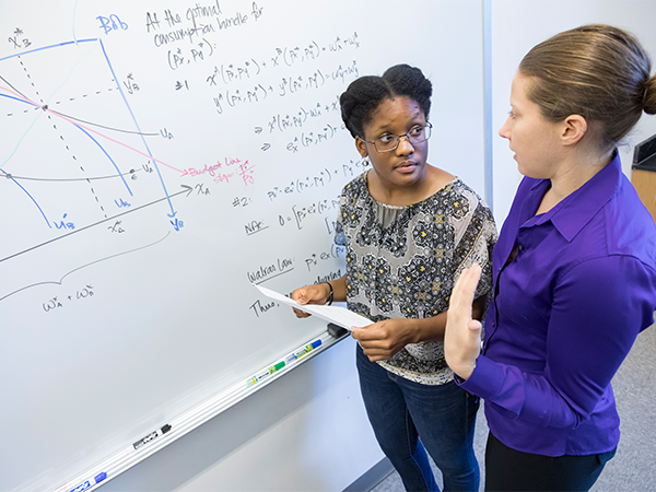 A student holding a paper talks with a female professor while they stand in front of a white board filled with formulas, graphs and data.